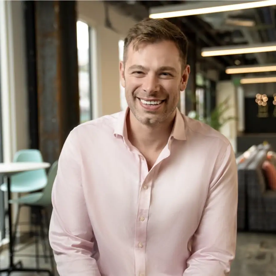 A man smiling in a pink shirt, looking toward camera.